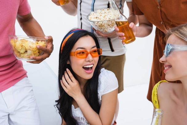 excité asiatique femme en lunettes de soleil rire près de amis avec des collations et de la bière - Photo, image