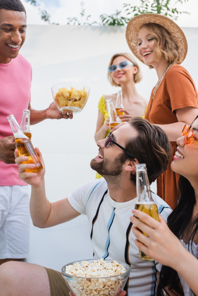 feliz interracial los hombres tintineo con botellas de cerveza cerca sonriendo las mujeres  - Foto, imagen