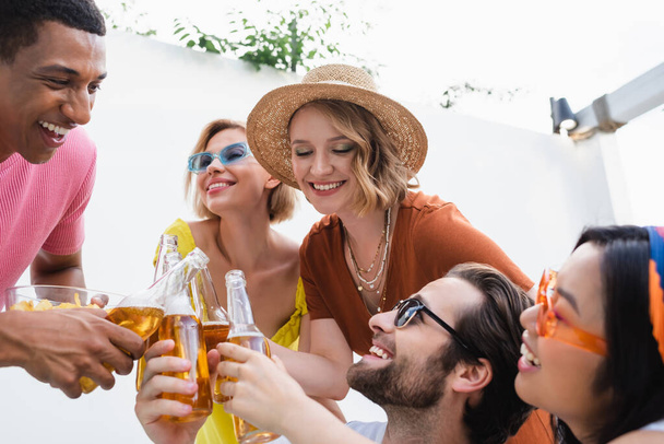 amigos multiculturales satisfechos tintinear botellas de cerveza durante la fiesta de verano al aire libre - Foto, Imagen