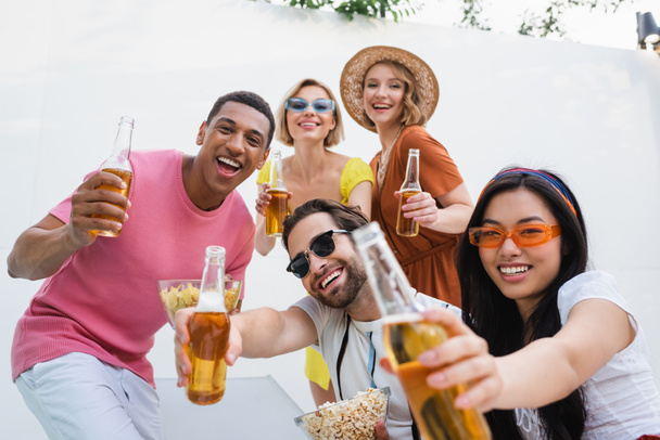 cheerful multiethnic friends toasting with beer while looking at camera during party - 写真・画像