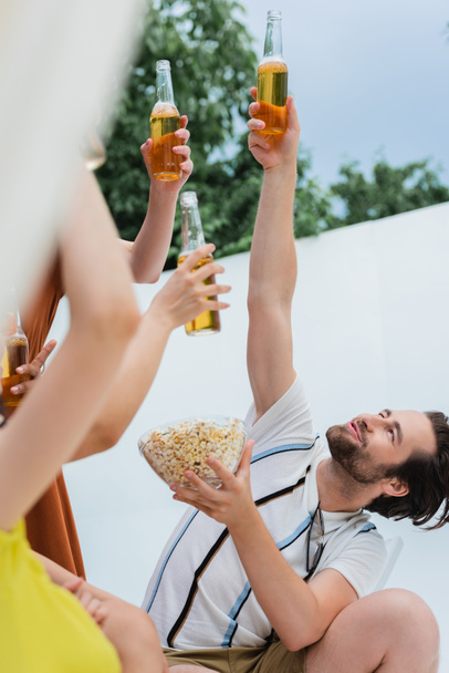 cheerful man with popcorn toasting with beer near women on blurred foreground - 写真・画像