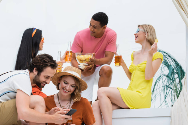 улыбающийся мужчина показывает мобильный телефон женщине в соломенной шляпе возле мультикультурных друзей с пивом - Фото, изображение