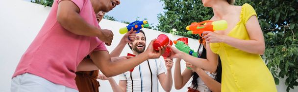 excited multicultural friends with water pistols toasting with plastic cups during party, banner - Fotoğraf, Görsel