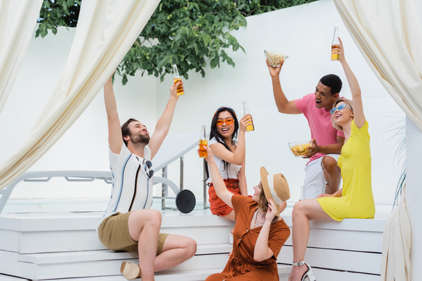 молодые и счастливые многонациональные друзья, пьющие пиво во время летней вечеринки в патио - Фото, изображение