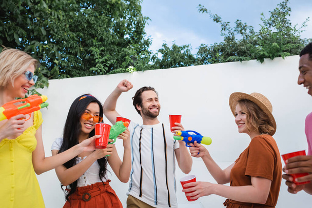 cheerful man showing win gesture near multicultural friends with water pistols and plastic cups - Photo, Image
