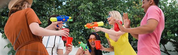 возбужденные женщины играют с водяными пистолетами во время летней вечеринки с многонациональными друзьями, баннер - Фото, изображение