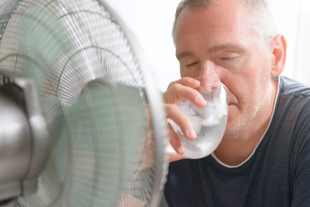 Mies kärsii lämpöä kotona juo kylmää vettä tai muuta juomaa ja yrittää jäähtyä tuuletin - Valokuva, kuva