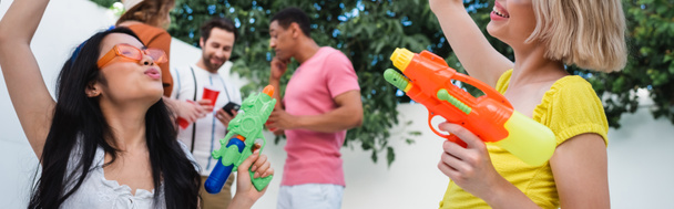 joyeux interracial femmes tenant pistolets à eau près des amis sur fond flou, bannière - Photo, image