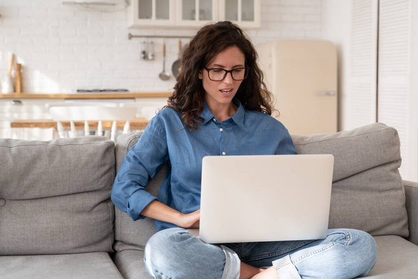 Frustrierte Frau schaut auf Laptop-Bildschirm mit offenem Mund und Misstrauen auf Gesicht, wenn sie zu Hause auf dem Sofa sitzt - Foto, Bild