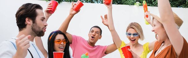 happy man showing win gesture near multicultural friends toasting with plastic cups during party, banner - Foto, imagen