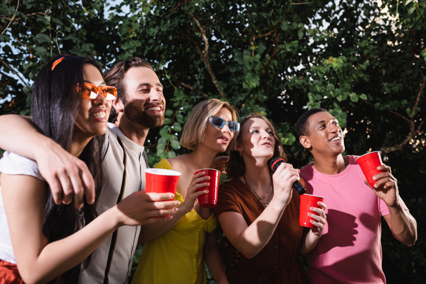 amigos multiétnicos alegres abraçando e cantando karaoke durante a festa de verão - Foto, Imagem