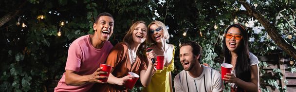 izgatott multikulturális barátok énekelnek karaoke a buli alatt az éjszakai kertben, banner - Fotó, kép