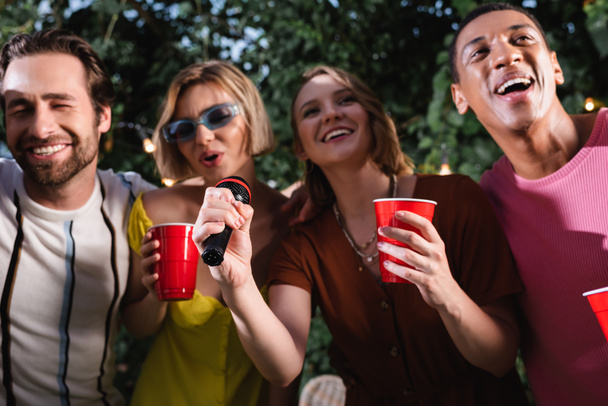 Cheerful multiethnic friends with plastic cups singing karaoke outdoors  - Foto, afbeelding