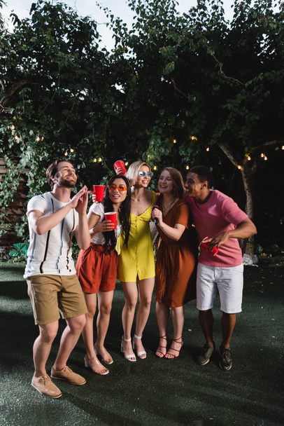 Молодые многонациональные друзья поют караоке и держат пластиковые стаканчики на траве вечером  - Фото, изображение