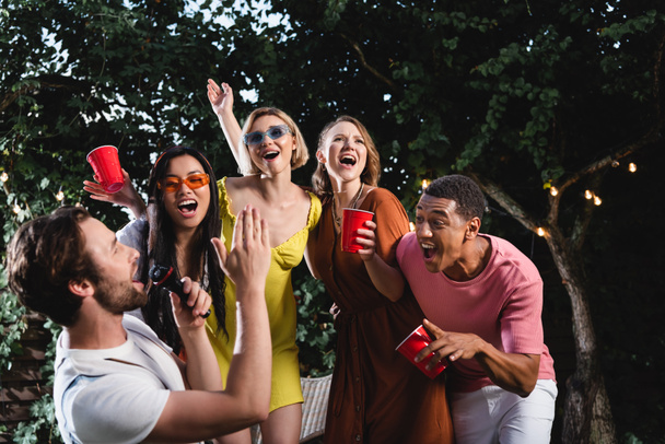 Man singing in microphone near positive multicultural friends with plastic cups outdoors in evening  - Photo, Image