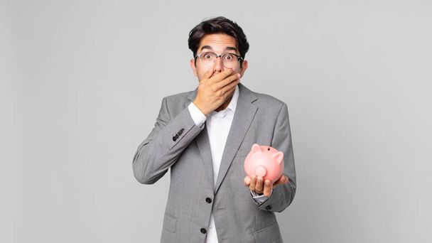 young hispanic man covering mouth with hands with a shocked and holding a piggy bank - 写真・画像