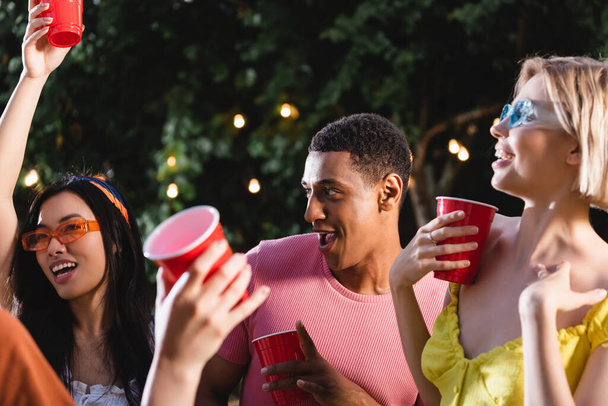 African american man holding plastic cup near interracial smiling friends outdoors  - Zdjęcie, obraz