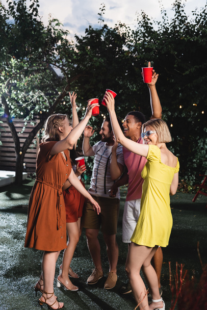 Ενθουσιασμένοι πολυεθνικοί άνθρωποι σηκώνουν πλαστικά ποτήρια κατά τη διάρκεια του πάρτι το βράδυ  - Φωτογραφία, εικόνα