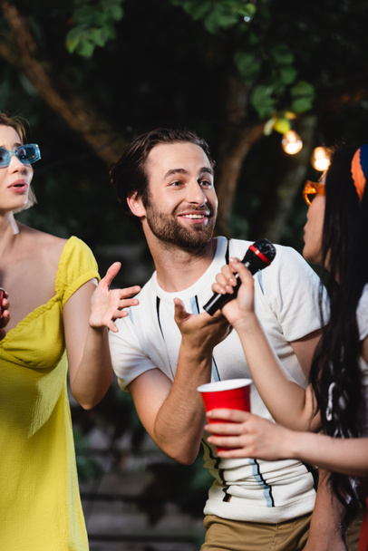 Young people pointing at asian friend singing karaoke and holding plastic cup  - Photo, Image