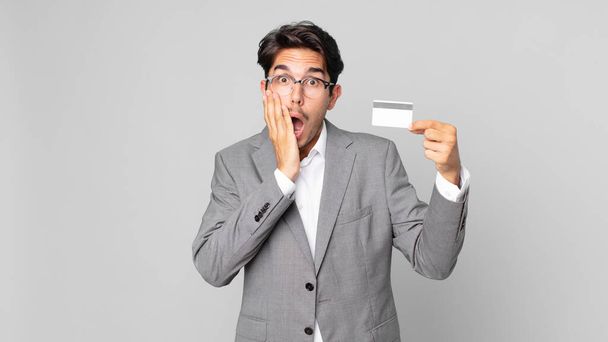 young hispanic man feeling shocked and scared and holding a credit card - Foto, imagen