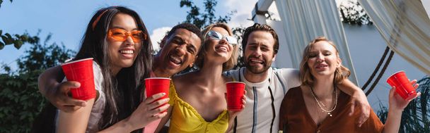 Amigos interracial positivos con copas de plástico abrazándose al aire libre, pancarta  - Foto, imagen