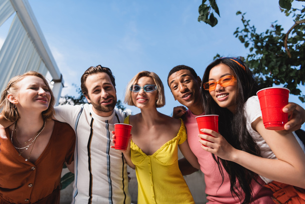 Vista ad angolo basso di amici multiculturali con drink guardando la fotocamera durante la festa  - Foto, immagini
