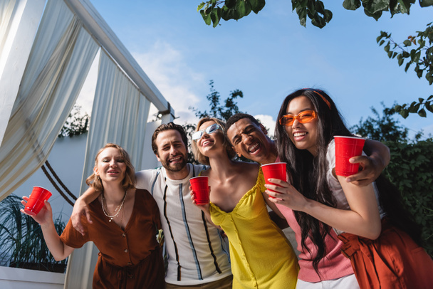 Feliz interracial de los jóvenes con vasos de plástico abrazando al aire libre  - Foto, imagen