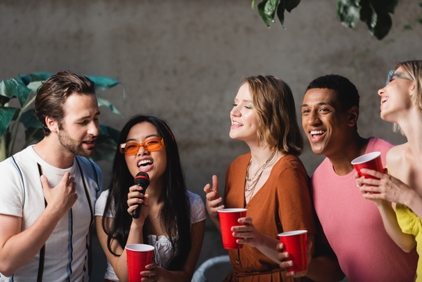 Asian woman with microphone singing near interracial friends holding plastic cups in evening  - Foto, afbeelding