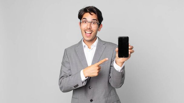 young hispanic man looking excited and surprised pointing to the side and holding a smartphone - Foto, Imagen
