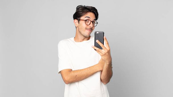young hispanic man shrugging, feeling confused and uncertain and holding a smartphone - Foto, immagini