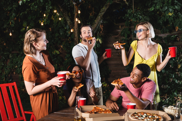 Felices amigos multiculturales comiendo pizza cerca de tazas de plástico y cerveza al aire libre  - Foto, Imagen