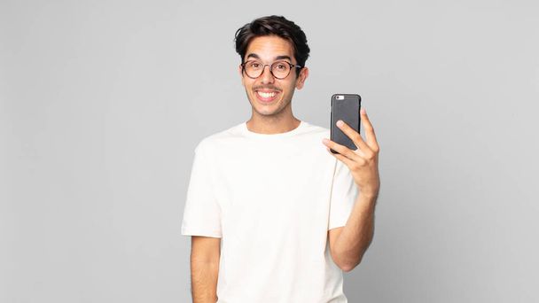 young hispanic man looking happy and pleasantly surprised and holding a smartphone - Φωτογραφία, εικόνα
