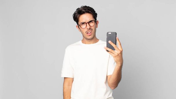 young hispanic man feeling puzzled and confused and holding a smartphone - Φωτογραφία, εικόνα