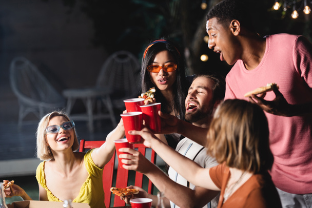 Excited interracial people toasting with plastic cups and holding pizza outdoors in evening  - Φωτογραφία, εικόνα
