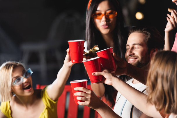 Plastic cups in hands of interracial friends toasting on blurred background  - Photo, Image