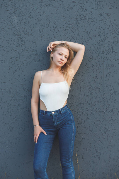 young blonde woman in the white sleeveless t-shirt and blue jeans standing over gray wall background - Zdjęcie, obraz