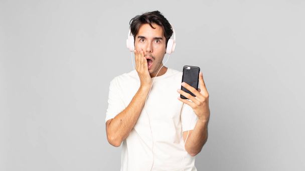 young hispanic man feeling shocked and scared with headphones and smartphone - Photo, Image