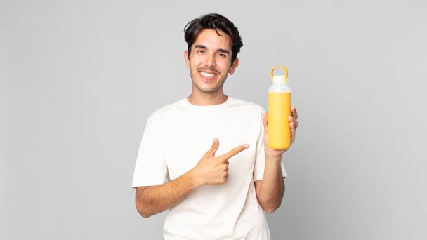 young hispanic man smiling cheerfully, feeling happy and pointing to the side with a coffee thermos - 写真・画像