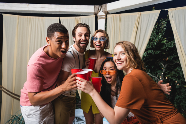 Gente interracial emocionada brindando con copas de plástico en el patio por la noche  - Foto, Imagen