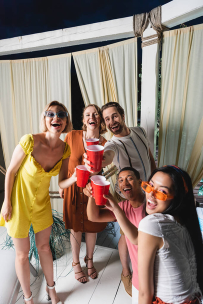 パティオでの夏のパーティー中にカメラを見てプラスチックカップを持つ積極的な多民族の人々 - 写真・画像