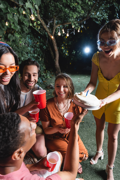 Femme tenant gâteau d'anniversaire près des amis interracial avec des tasses en plastique à l'extérieur  - Photo, image
