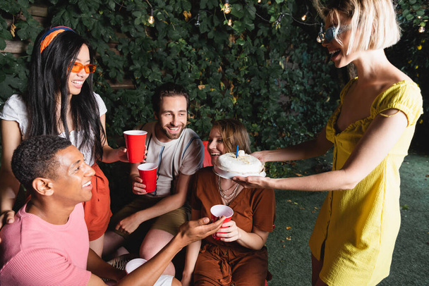 Woman holding birthday cake near african american friend and people with plastic cups outdoors  - Photo, Image