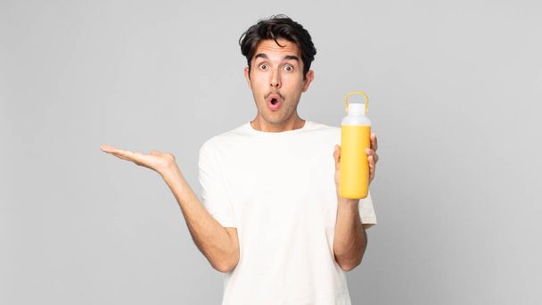 young hispanic man looking surprised and shocked, with jaw dropped holding an object with a coffee thermos - Foto, Imagem