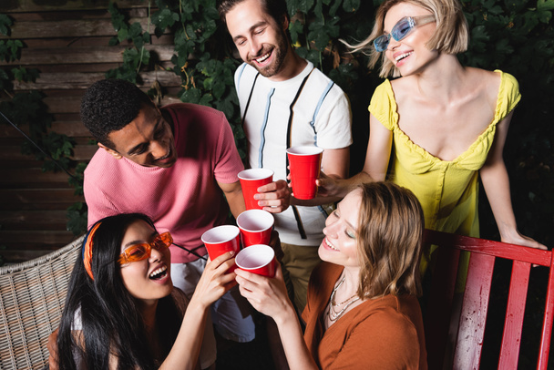 Cheerful multicultural people toasting with plastic cups outdoors at night  - Photo, Image