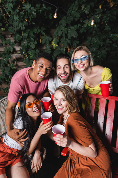 Smiling interracial friends with plastic cups hugging and looking at camera outdoors  - Foto, imagen
