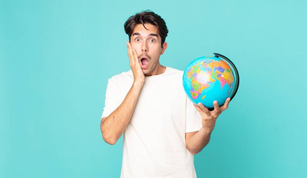 young hispanic man feeling shocked and scared and holding a world globe map - Foto, immagini