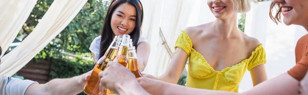 Hymyilevä aasialainen nainen paahtaminen oluen kanssa lähellä ystäviä ulkona, banneri  - Valokuva, kuva