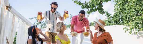 Smiling interracial men holding snacks and beer near friends in patio, banner  - Photo, Image