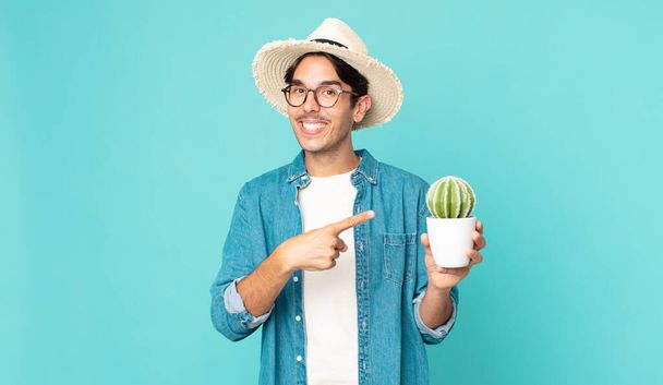 young hispanic man looking excited and surprised pointing to the side and holding a cactus - Фото, изображение