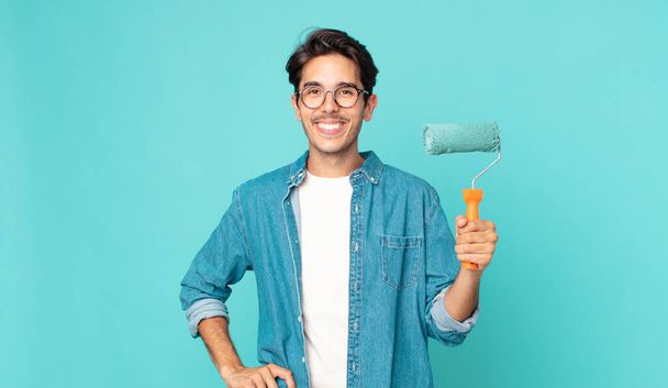 young hispanic man smiling happily with a hand on hip and confident and holding a paint roller - 写真・画像
